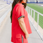 Woman with dark hair standing in a side profile position wearing the Daisy Top and Jane Skirt at a football field. The focal point of this image is the striping down side of the skirt as well as around the cuff. 