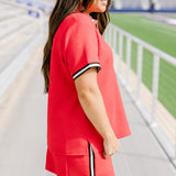 Woman with dark hair standing in a side profile position wearing the Daisy Top and Jane Skirt at a football field. The focal point of this image is the striping down side of the skirt as well as around the cuff. 