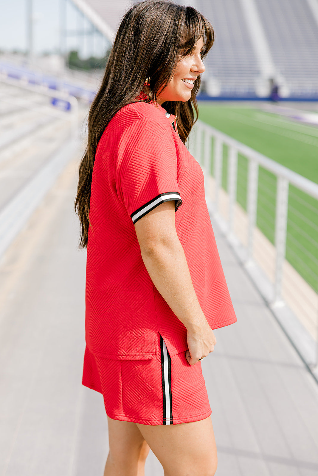 Woman with dark hair standing in a side profile position wearing the Daisy Top and Jane Skirt at a football field. The focal point of this image is the striping down side of the skirt as well as around the cuff. 