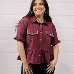 A woman wearing a short-sleeve button-up shirt with vertical stripes in burgundy and dark red, paired with dark blue jeans, standing against a white wood-paneled wall.