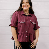A woman wearing a short-sleeve button-up shirt with vertical stripes in burgundy and dark red, paired with dark blue jeans, standing against a white wood-paneled wall.