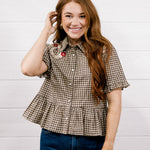 A smiling woman with long red hair models a brown and beige houndstooth short-sleeve button-up shirt with a ruffled hem, paired with blue jeans, against a white wood-paneled wall.