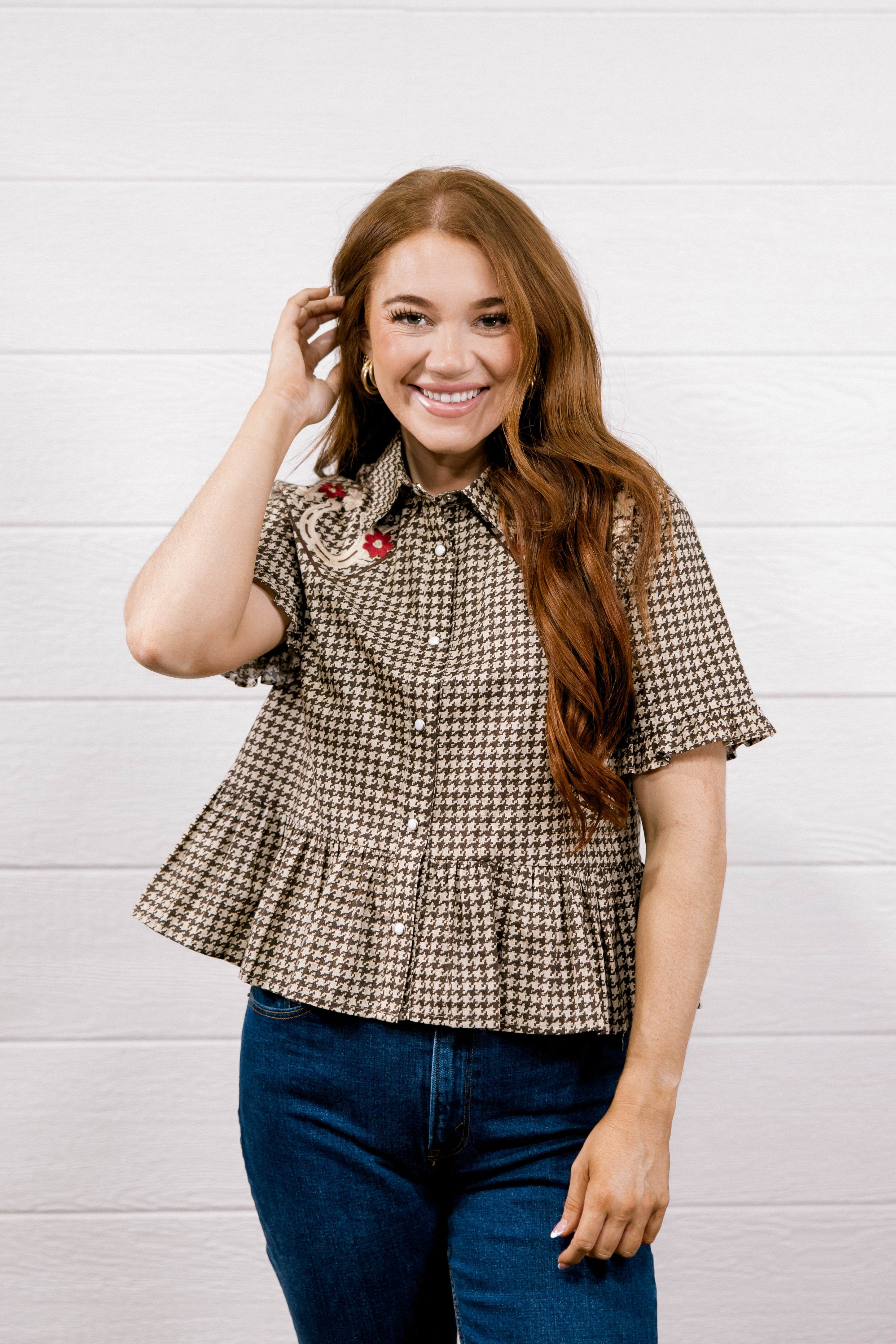 A smiling woman with long red hair models a brown and beige houndstooth short-sleeve button-up shirt with a ruffled hem, paired with blue jeans, against a white wood-paneled wall.