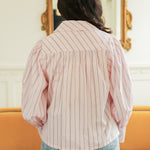 Woman with dark hair standing in a back profile position wearing the Dolly Top in a lifestyle scene. The focal point of this image is the pink and brown striped pattern on back as well as long sleeves. The model paired this top with light-wash denim jeans. 