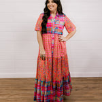 Woman wearing a colorful patterned dress standing against a white wooden panel background facing the front. 