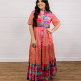Woman wearing a colorful patterned dress standing against a white wooden panel background facing the front. 