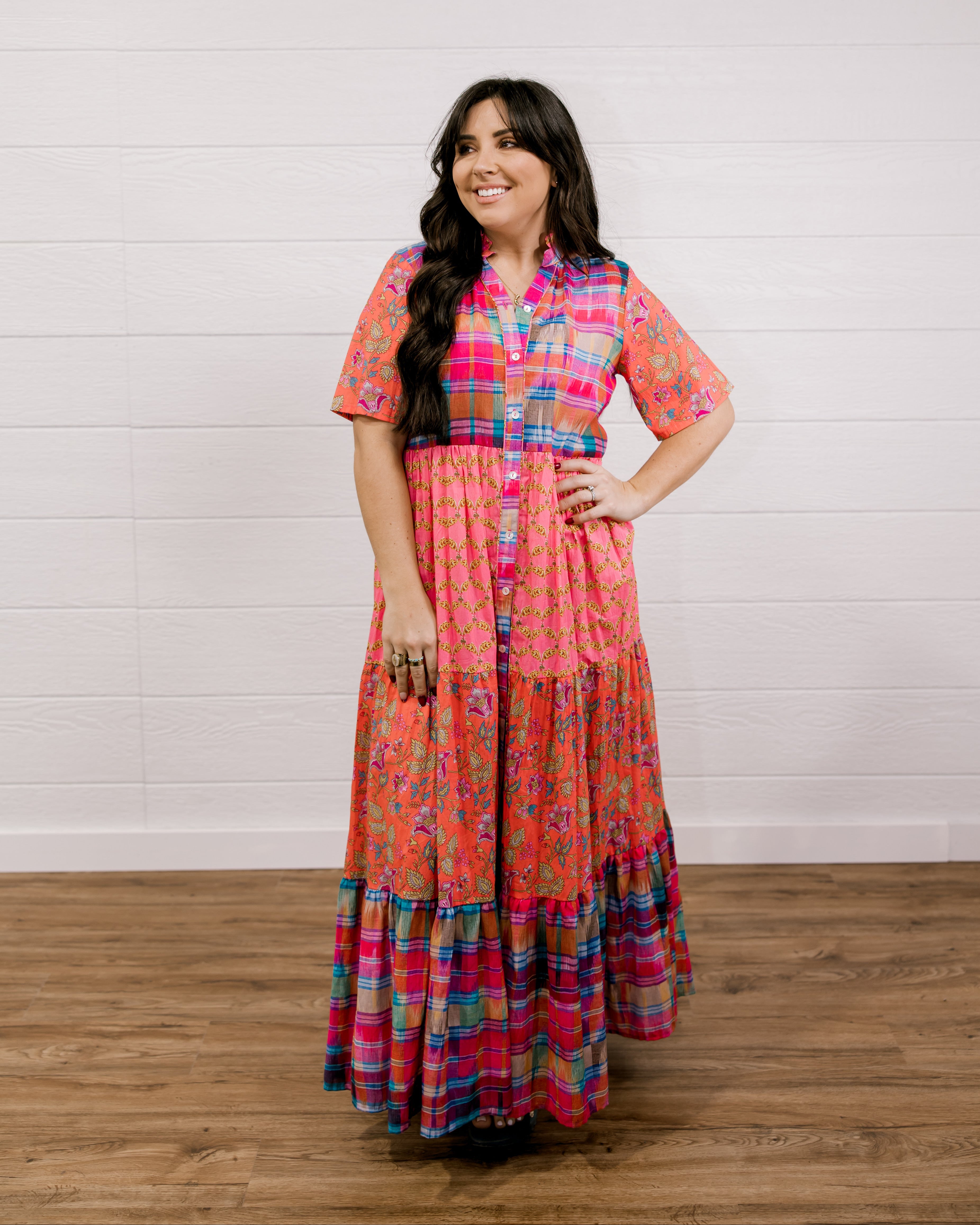 Woman wearing a colorful patterned dress standing against a white wooden panel background facing the front. 