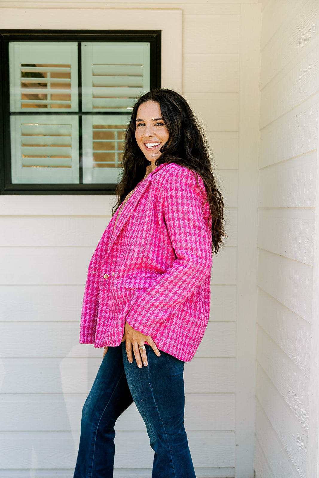 Woman with dark hair wearing a pink tweed blazer with gold buttons. Model paired blazer with dark-wash denim jeans. 