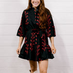 A woman modeling a black mini dress with red floral embroidery and white lace cowboy boots, standing against a white shiplap wall and wood floor.