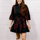 A woman modeling a black mini dress with red floral embroidery and white lace cowboy boots, standing against a white shiplap wall and wood floor.