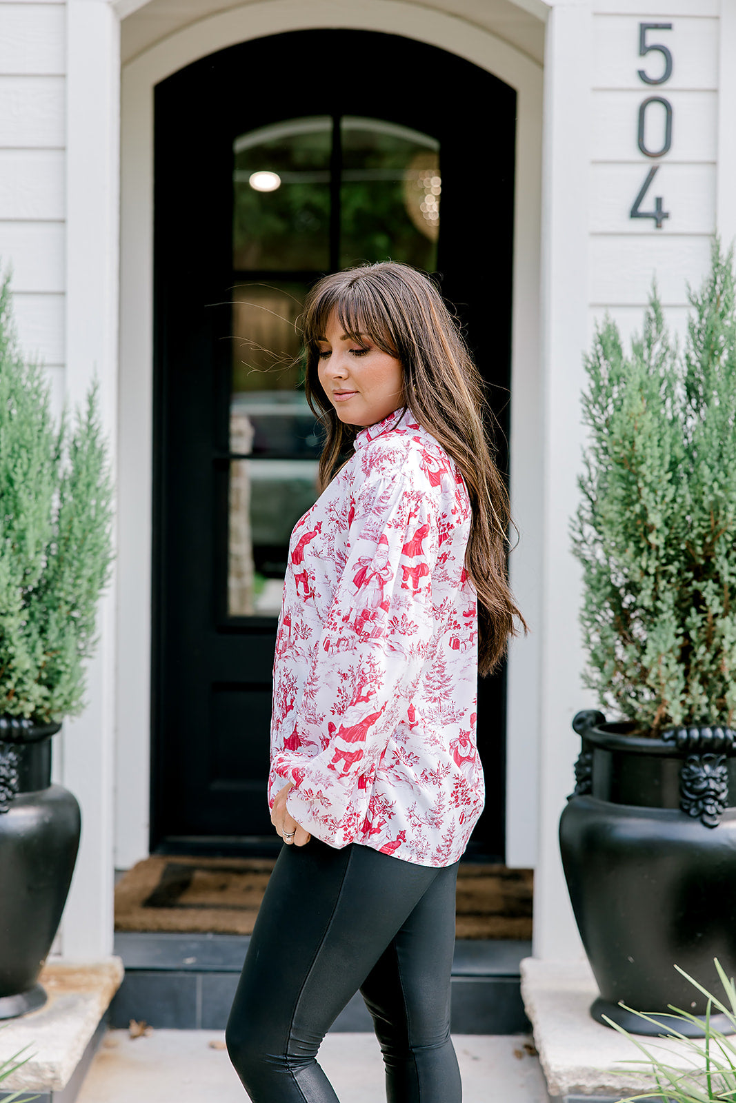 Woman with dark hair wearing a long-sleeve red & cream Santa toile graphics top. Model paired this top with leather pants. 