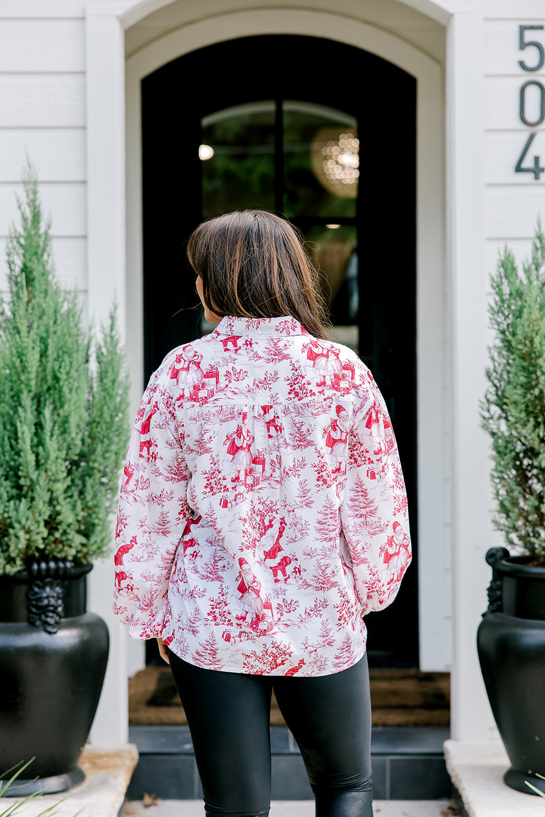 Woman with dark hair wearing a long-sleeve red & cream Santa toile graphics top. Model paired this top with leather pants. 