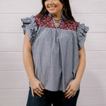 A woman modeling a short-sleeve blouse with ruffled cap sleeves, featuring a blue and white vertical stripe body and a red and white floral patterned yoke, paired with dark jeans against a white wood-paneled wall.