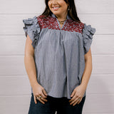 A woman modeling a short-sleeve blouse with ruffled cap sleeves, featuring a blue and white vertical stripe body and a red and white floral patterned yoke, paired with dark jeans against a white wood-paneled wall.
