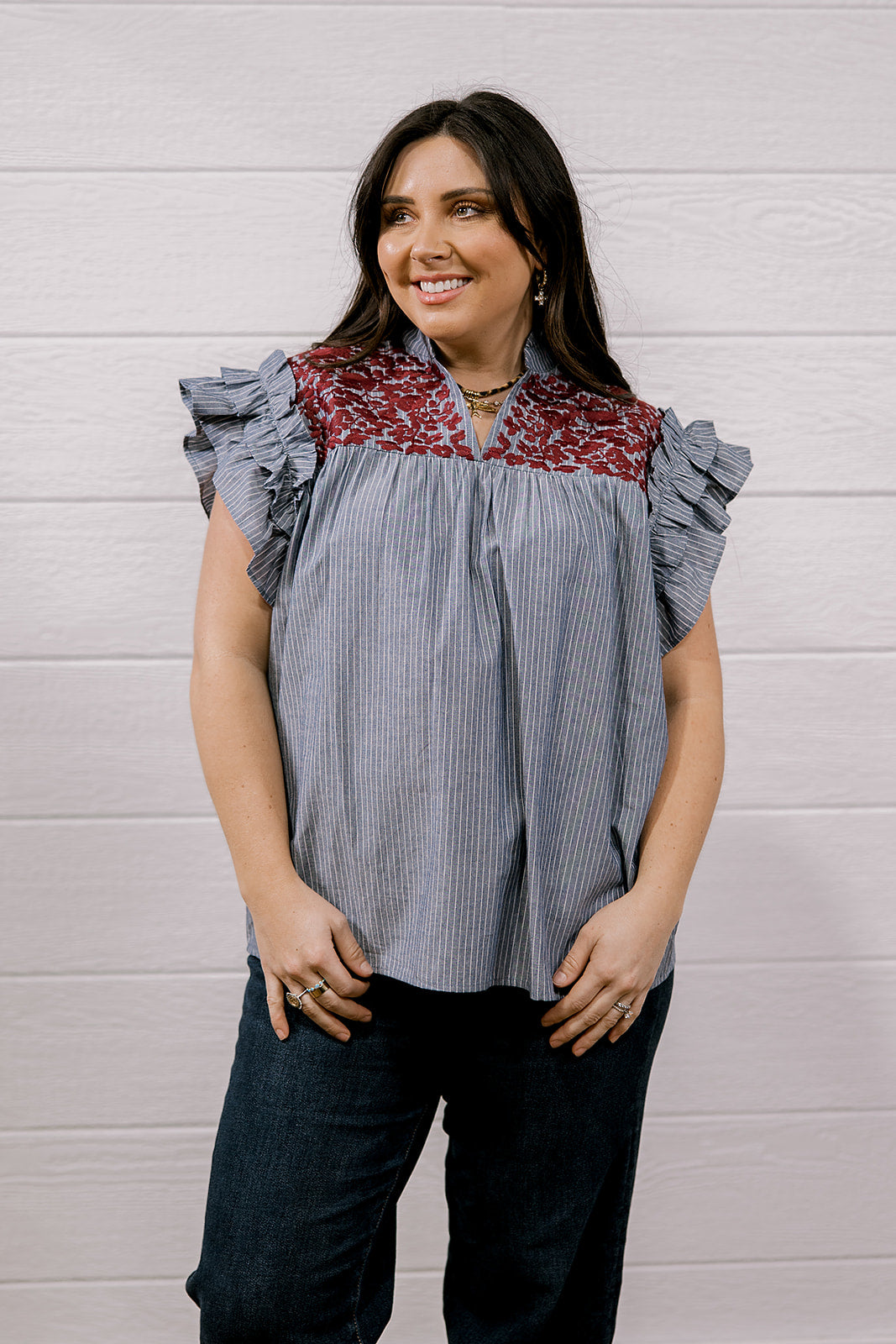 A woman modeling a short-sleeve blouse with ruffled cap sleeves, featuring a blue and white vertical stripe body and a red and white floral patterned yoke, paired with dark jeans against a white wood-paneled wall.