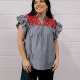 A woman wearing a gray and white vertically striped blouse with a red floral yoke and ruffled cap sleeves, paired with dark jeans, standing against a white wood-paneled wall.