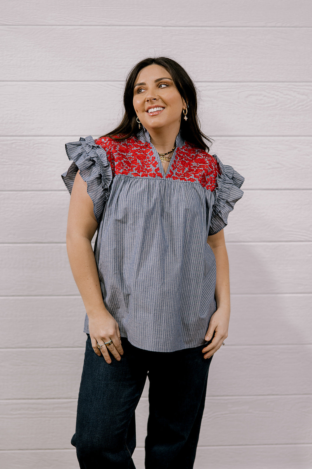 A woman wearing a gray and white vertically striped blouse with a red floral yoke and ruffled cap sleeves, paired with dark jeans, standing against a white wood-paneled wall.