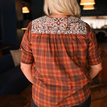 Woman with blonde hair wearing a short-sleeve top with brown plaid graphics, and white embroidery across shoulders and chest. Model paired this top with dark-wash denim jeans. 
