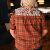 Woman with blonde hair wearing a short-sleeve top with brown plaid graphics, and white embroidery across shoulders and chest. Model paired this top with dark-wash denim jeans. 