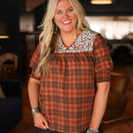 Woman with blonde hair wearing a short-sleeve top with brown plaid graphics, and white embroidery across shoulders and chest. Model paired this top with dark-wash denim jeans. 