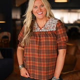 Woman with blonde hair wearing a short-sleeve top with brown plaid graphics, and white embroidery across shoulders and chest. Model paired this top with dark-wash denim jeans. 