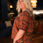 Woman with blonde hair wearing a short-sleeve top with brown plaid graphics, and white embroidery across shoulders and chest. Model paired this top with dark-wash denim jeans. 