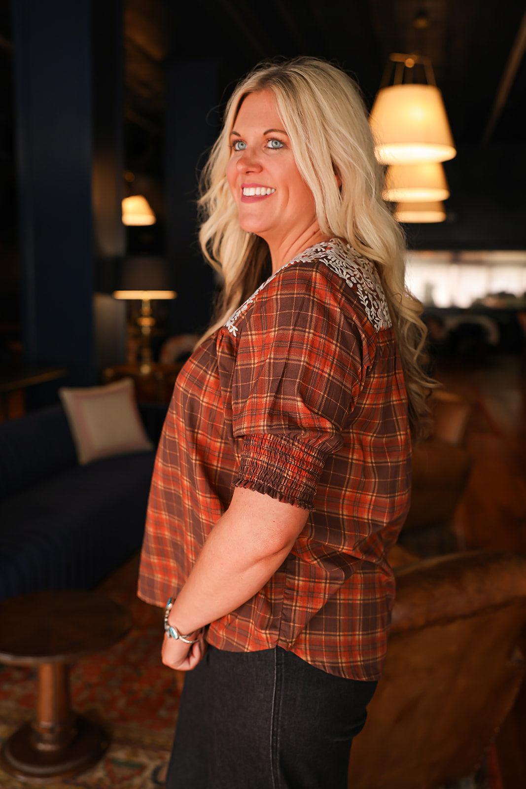 Woman with blonde hair wearing a short-sleeve top with brown plaid graphics, and white embroidery across shoulders and chest. Model paired this top with dark-wash denim jeans. 