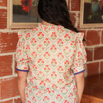 Woman with dark hair standing in a back profile position wearing the Haley Top against a red brick background. The focal point of this image is the red floral pattern and blue trimming on sleeves. The model paired this top with light-wash denim jeans. 