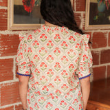 Woman with dark hair standing in a back profile position wearing the Haley Top against a red brick background. The focal point of this image is the red floral pattern and blue trimming on sleeves. The model paired this top with light-wash denim jeans. 