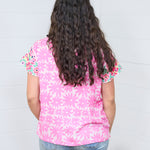 Woman with dark hair standing in a back profile position wearing the Hallie Top against a white background. The focal point of this image is the graphics on the back of the top with emphasis on the embroidery on the back of the sleeves. The model paired this top with light-wash denim jeans.