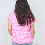 Woman with dark hair standing in a back profile position wearing the Hallie Top against a white background. The focal point of this image is the graphics on the back of the top with emphasis on the embroidery on the back of the sleeves. The model paired this top with light-wash denim jeans.