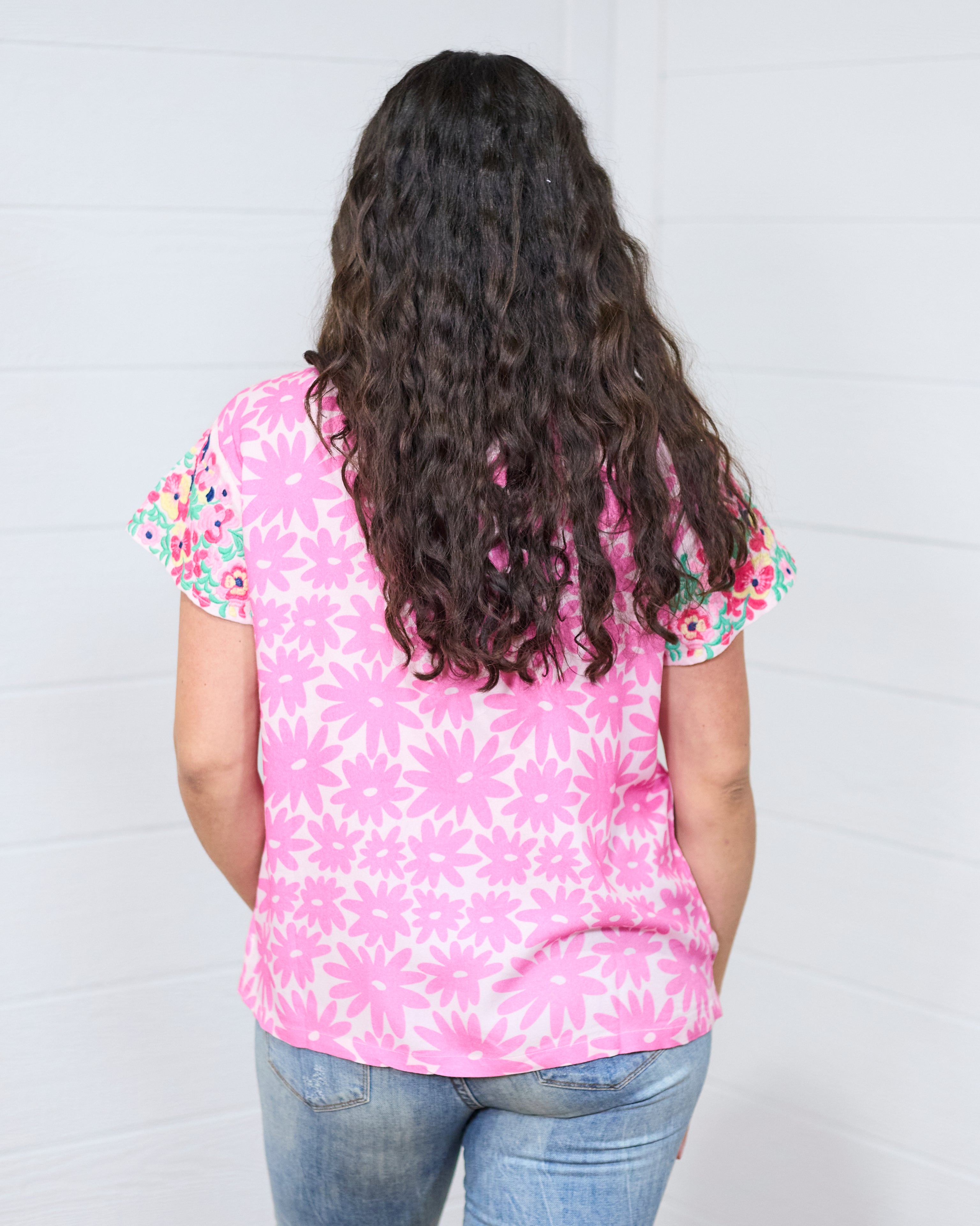 Woman with dark hair standing in a back profile position wearing the Hallie Top against a white background. The focal point of this image is the graphics on the back of the top with emphasis on the embroidery on the back of the sleeves. The model paired this top with light-wash denim jeans.