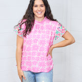 Woman with dark hair standing in a front profile position wearing the Hallie Top against a white background. The focal point of this image is the front graphics with emphasis on high pleated neckline and embroidery on front of sleeves. The model paired this top with light-wash denim jeans. 