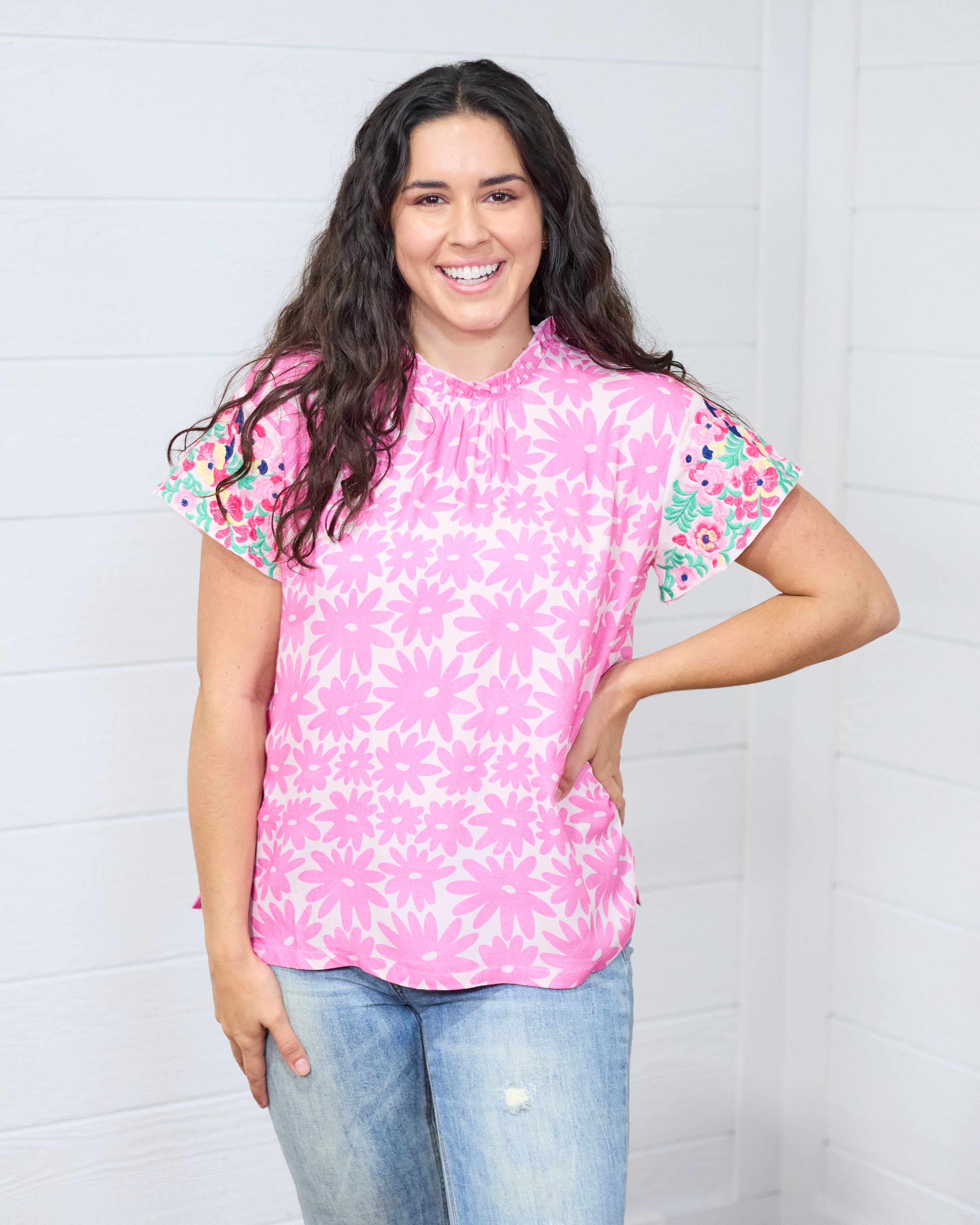 Woman with dark hair standing in a front profile position wearing the Hallie Top against a white background. The focal point of this image is the front graphics with emphasis on high pleated neckline and embroidery on front of sleeves. The model paired this top with light-wash denim jeans. 