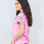 Woman with dark hair standing in a side profile position wearing the Hallie Top standing against a white background. The focal point of this image is the embroidery on the sleeves and graphics on the side of the top. The model paired this top with light-wash denim jeans.