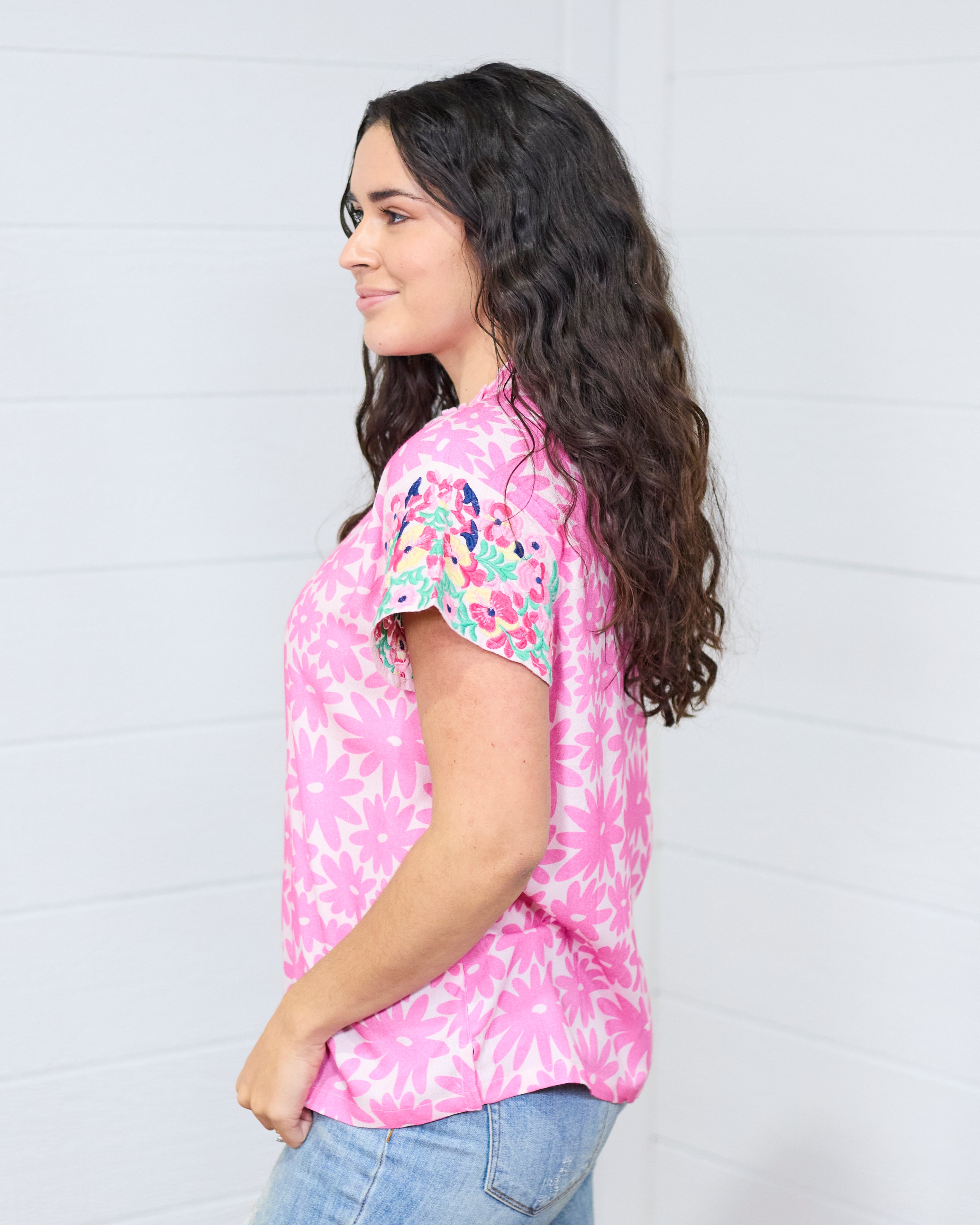 Woman with dark hair standing in a side profile position wearing the Hallie Top standing against a white background. The focal point of this image is the embroidery on the sleeves and graphics on the side of the top. The model paired this top with light-wash denim jeans.
