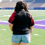 Woman with dark hair wearing a short-sleeve black top with red floral embroidery on sleeves. The model paired this top with light-wash denim shorts. 