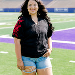 Woman with dark hair wearing a short-sleeve black shirt with red floral embroidery on the sleeves. The model paired this top with light-wash denim shorts. 