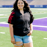 Woman with dark hair wearing a short-sleeve black shirt with red floral embroidery on the sleeves. The model paired this top with light-wash denim shorts. 