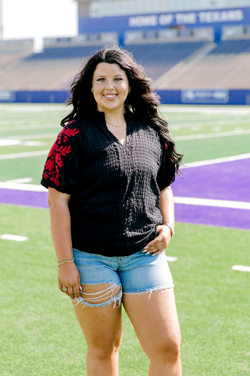Woman with dark hair wearing a short-sleeve black shirt with red floral embroidery on the sleeves. The model paired this top with light-wash denim shorts. 