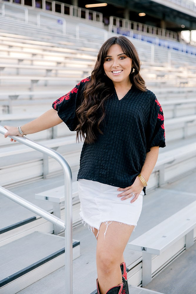 Woman with dark hair wearing a short-sleeve black top with red floral embroidery. The model paired this top with a white denim skirt. 