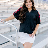 Woman with dark hair wearing a short-sleeve black top with red floral embroidery. The model paired this top with a white denim skirt. 
