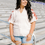 Woman with dark hair wearing a cream short-sleeve top with burnt orange embroidery, and light-wash denim shorts.