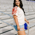 Woman with dark hair standing in a side profile position wearing the Havanah Top at a football field. The focal point of this image is the ruffled sleeves with burnt orange floral embroidery. The model paired this top with light-wash denim shorts.