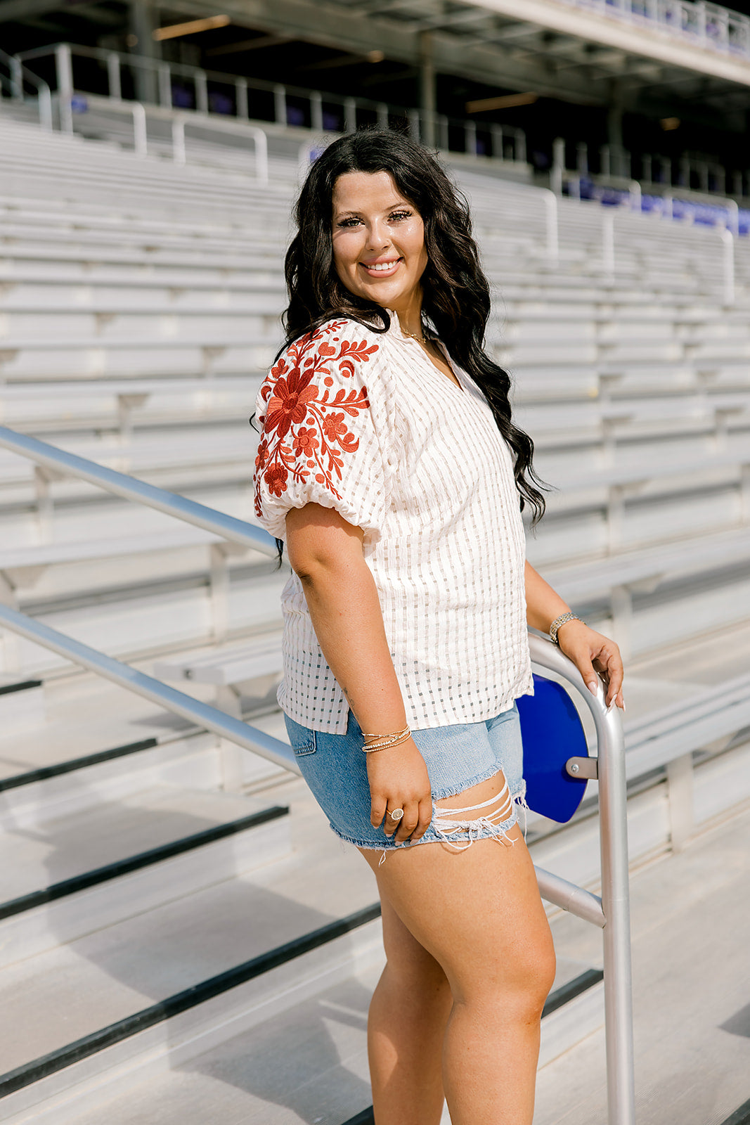 Woman with dark hair standing in a side profile position wearing the Havanah Top at a football field. The focal point of this image is the ruffled sleeves with burnt orange floral embroidery. The model paired this top with light-wash denim shorts.