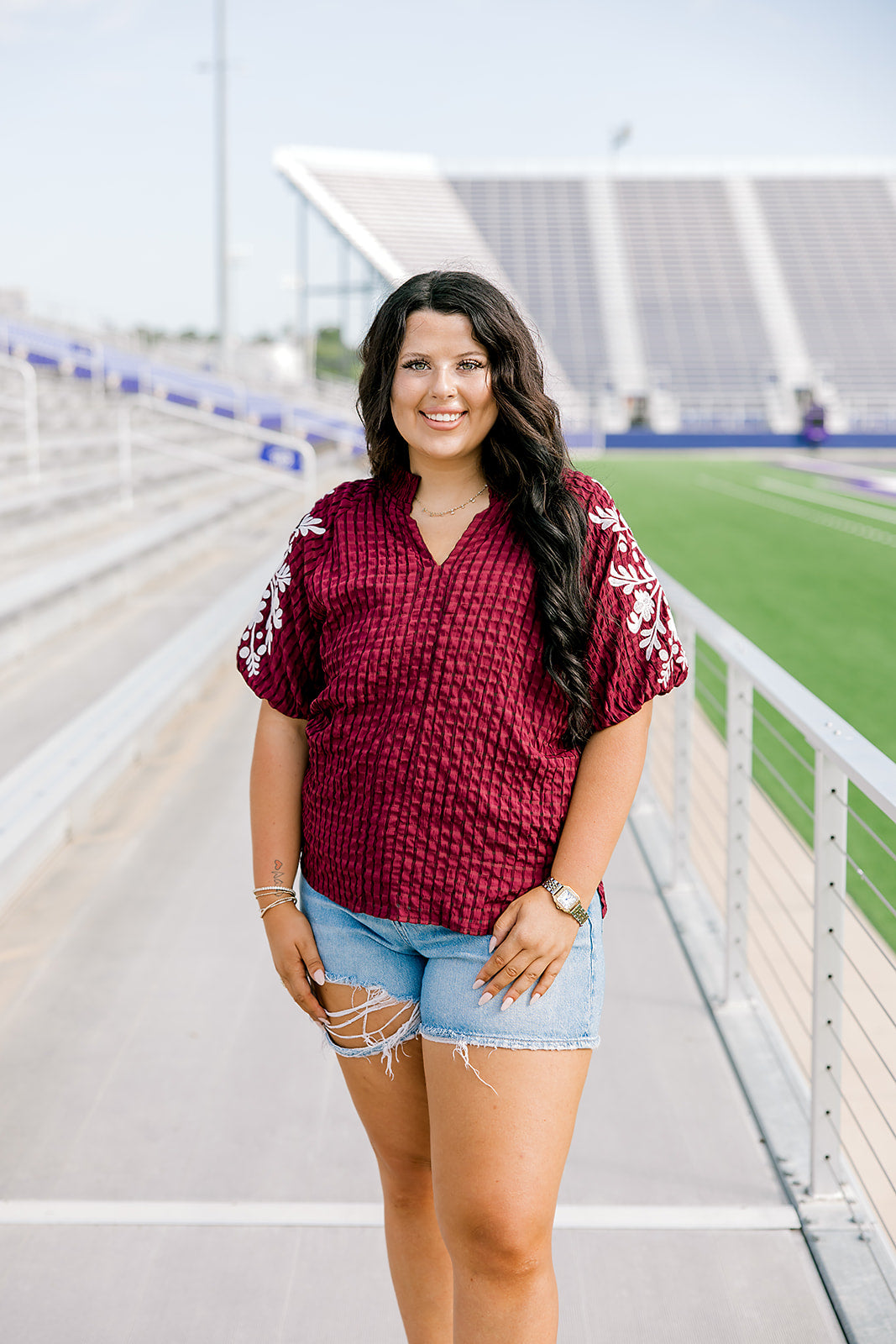 Woman with dark hair standing in a front profile position wearing the Havanah Top at a football field. The focal point of this image is the front details of the top including embroidery down shoulders, high-collar, and v-neckline. The model paired this top with light-wash denim shorts. 