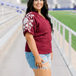 Woman with dark hair standing in a side profile position wearing the Havanah Top at a football field. The focal point of this image is the embroidery down the sleeve as well as the ruffle towards the cuff. The model paired this top with light-wash denim shorts.