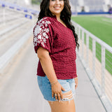 Woman with dark hair standing in a side profile position wearing the Havanah Top at a football field. The focal point of this image is the embroidery down the sleeve as well as the ruffle towards the cuff. The model paired this top with light-wash denim shorts.