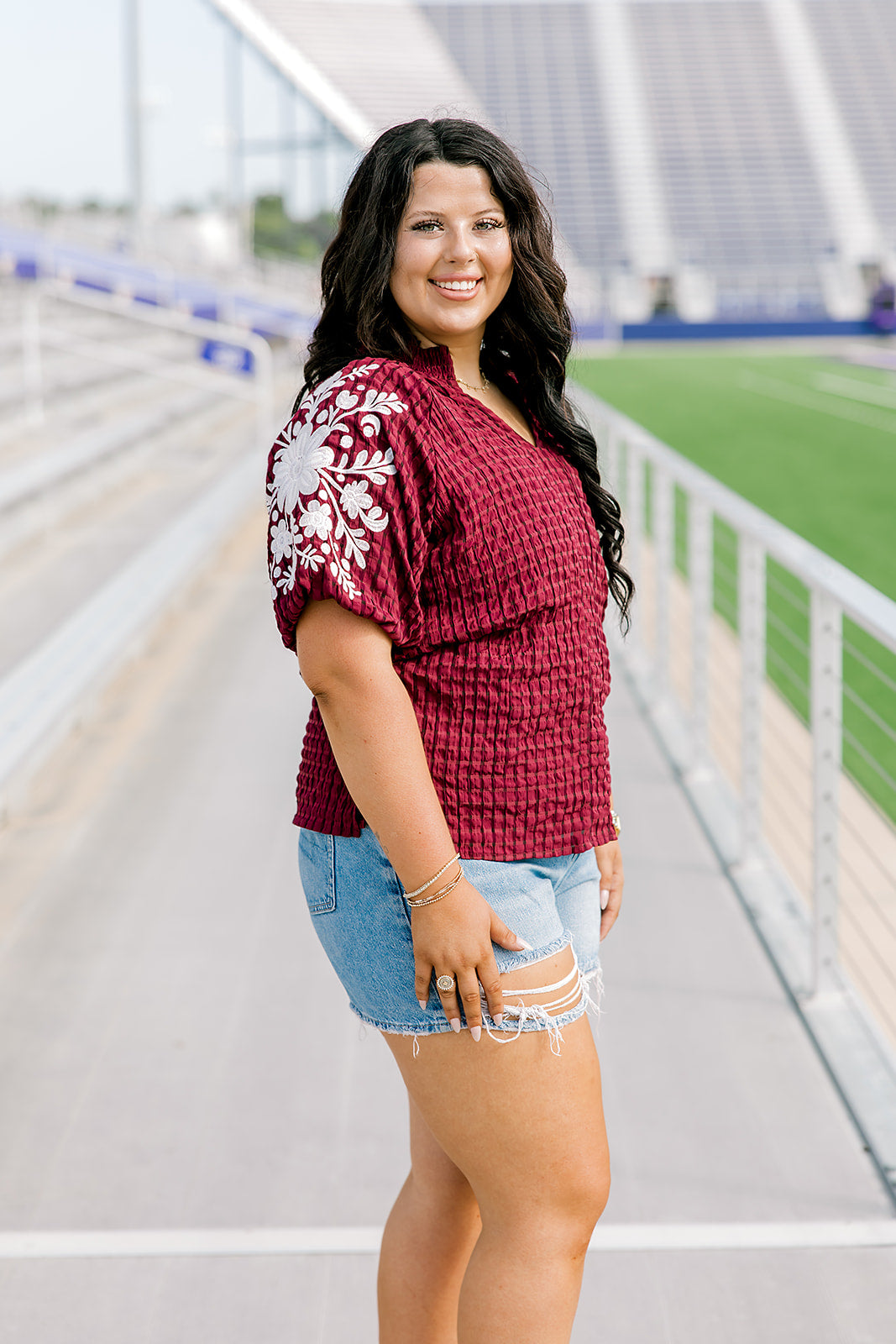 Woman with dark hair standing in a side profile position wearing the Havanah Top at a football field. The focal point of this image is the embroidery down the sleeve as well as the ruffle towards the cuff. The model paired this top with light-wash denim shorts.