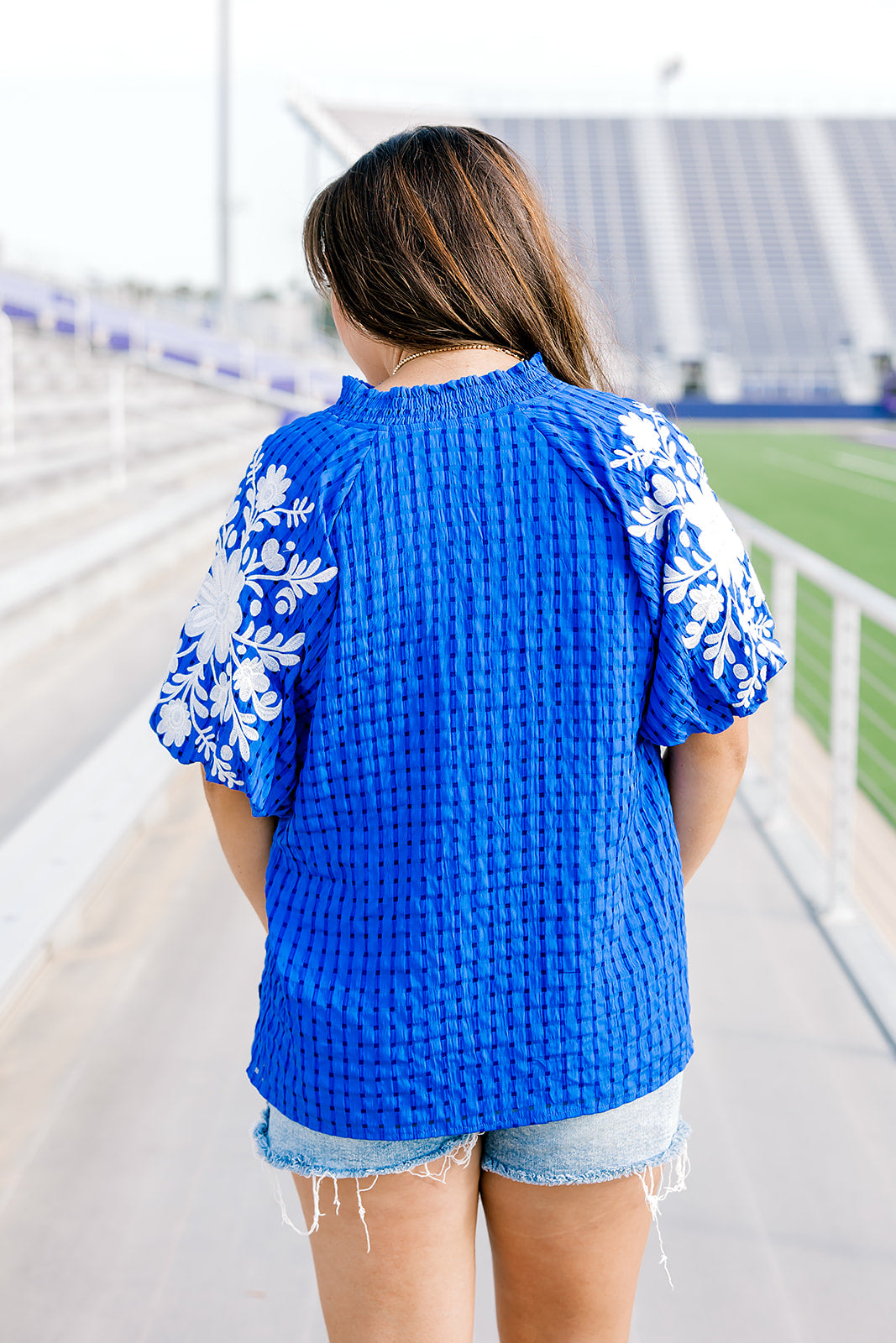 Woman with dark hair standing in a back profile position wearing the Havanah Top in a football field setting. The focal point of this image is the fabric material and white embroidery on the sleeves with slight emphasis on the roused neckline. The model paired these with light-wash denim shorts.